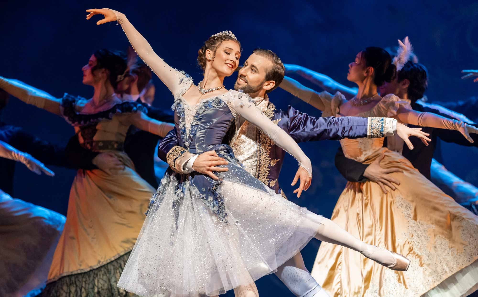 A ballerina in a sparkling blue costume is gracefully lifted by her partner in a matching suit, surrounded by dancers in elegant dresses, performing on stage under dramatic lighting.